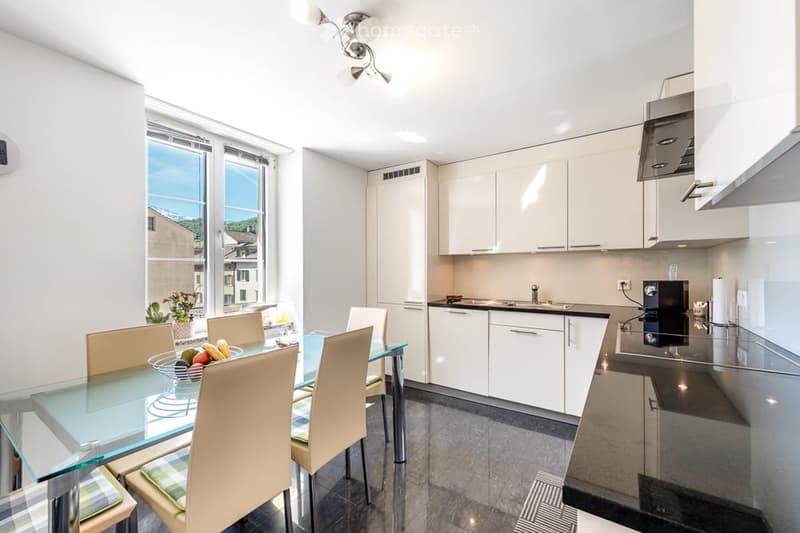 Modern open-plan kitchen and dining area with white cabinets, stainless steel appliances, a glass dining table with four chairs, and a fruit bowl on the table. Large windows offer a scenic view.