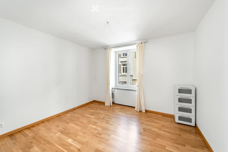 Empty room with wooden flooring, white walls, a window with curtains, a radiator, and a small cabinet with drawers.