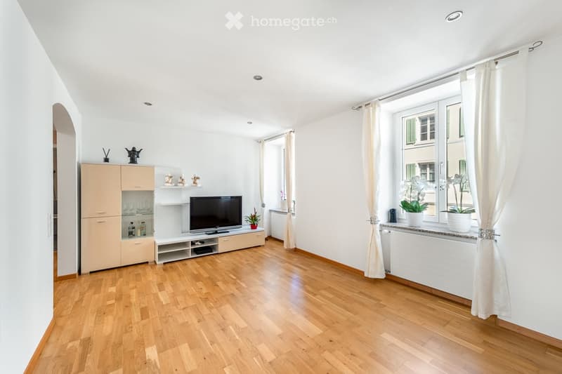 Bright living room with wooden flooring, large windows with white curtains, television on a stand, cabinet with glass doors, shelves with decorative items, and flower pots on the windowsill.