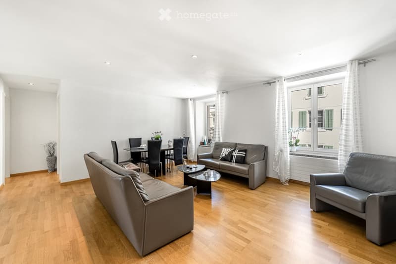 Modern living and dining area with hardwood flooring, grey leather sofas, coffee table, dining table with chairs, and decorative vases.