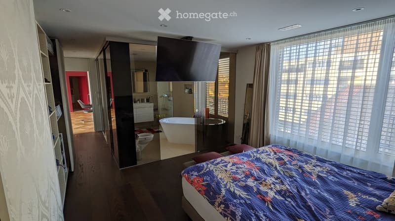 Modern bedroom with a large bed, a flat-screen TV, a large window with white curtains, and an en-suite bathroom with a bathtub and toilet.