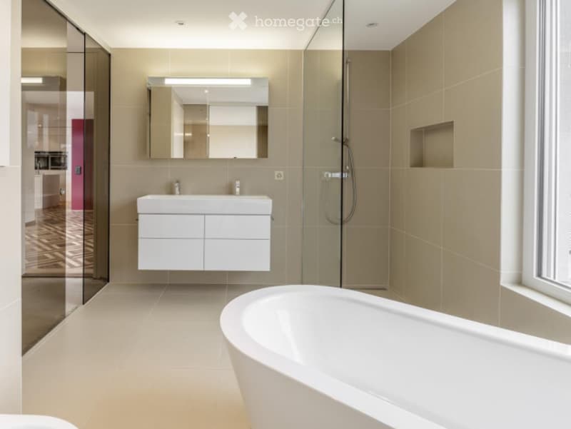 Modern bathroom with a freestanding bathtub, a white sink with a built-in cabinet, and a large mirror above it. The space features light beige tiles and a glass-enclosed shower area.