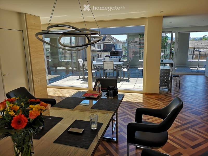 Modern living room with wooden herringbone flooring, large glass doors leading to a balcony, a sleek coffee table with a flower arrangement, and contemporary chairs.