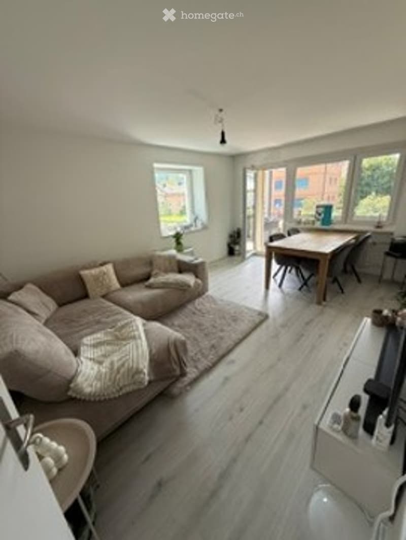 Bright, modern living and dining area with light wood flooring, large windows, a sectional sofa, coffee table, dining table with chairs, and a TV stand.