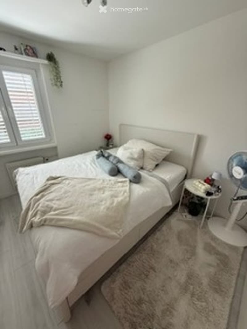 Bright bedroom with white walls, wooden flooring, large window with blinds, bed with pillows and blanket, nightstand with lamp, and a fan near the window.
