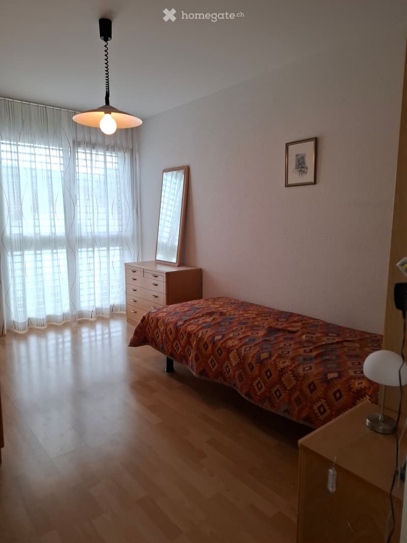 Cozy bedroom with wooden flooring, a bed with a colorful patterned bedspread, a nightstand with a lamp, a dresser with a mirror, and a hanging pendant light.