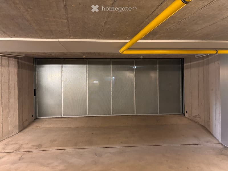 Empty underground parking space with concrete walls, a large metal door, and a yellow safety pipe running along the ceiling.