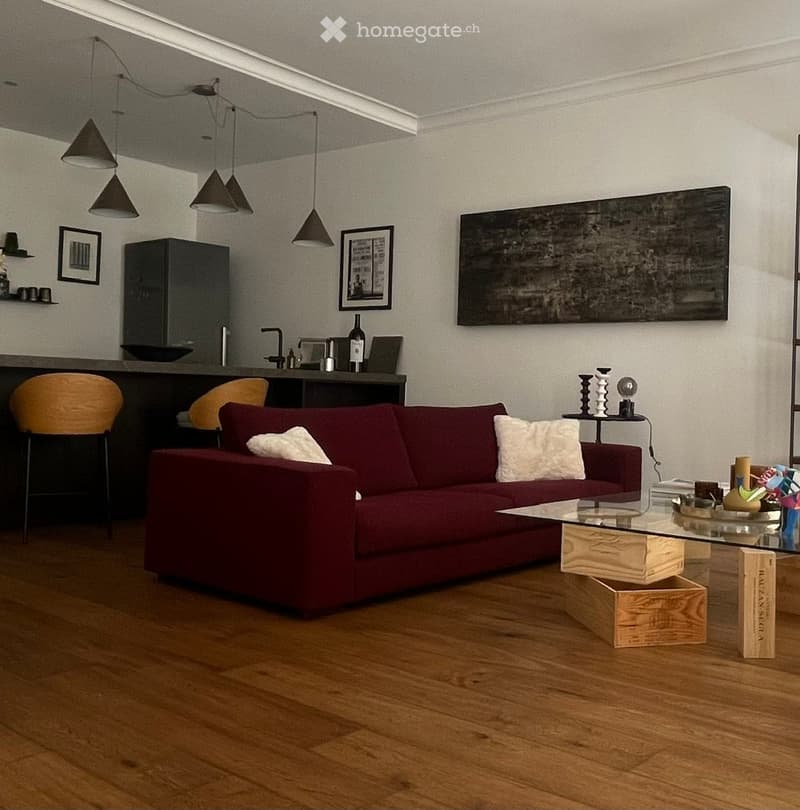 Modern living room with a red sofa, glass coffee table, pendant lights, and a small bar area with stools and a cabinet.