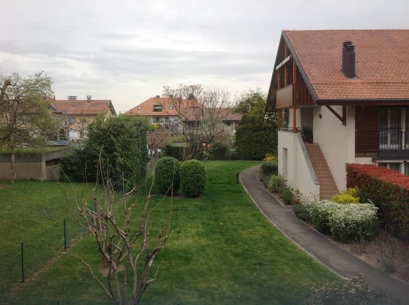 Well-maintained garden with green lawn, trimmed bushes, small tree, and pathway leading to a house with a wooden balcony and staircase.