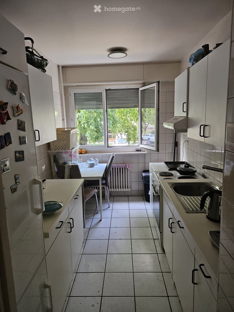 A compact kitchen with white tiled floors, white cabinets, a sink, stove, and a small dining area with a table and chairs. The kitchen has a window with blinds and a view of greenery outside.