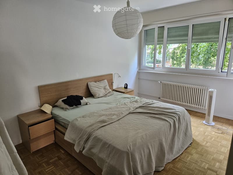 Cozy bedroom with a wooden bed, nightstands, a fan, and large windows with blinds. The room features a light-colored duvet and pillows, a wall-mounted lamp, and a radiator below the windows.
