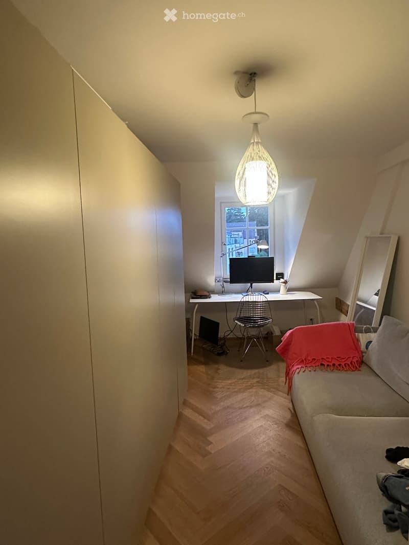 Cozy attic room with slanted ceilings, wooden flooring, a sofa with a pink throw, a desk with a laptop and chair, and a hanging pendant light.