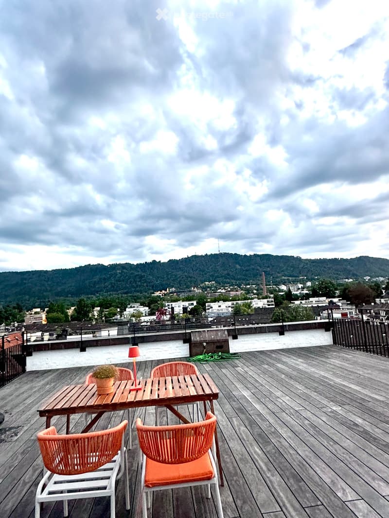 Wooden deck with a table, chairs, a lamp, and a small potted plant, overlooking a cityscape with hills in the background.
