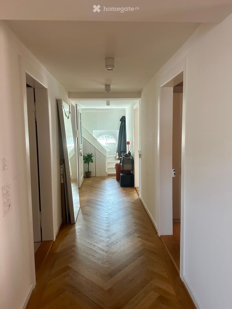 A long, well-lit hallway with wooden flooring and white walls, featuring several open doors leading to other rooms.
