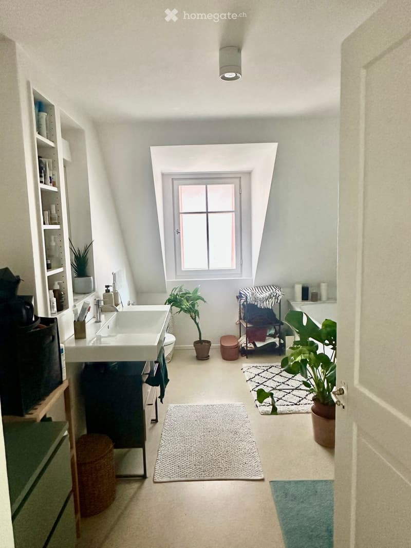Modern bathroom with a sink, toilet, bathtub, and potted plants. Features a large window, white walls, and multiple rugs on the floor.