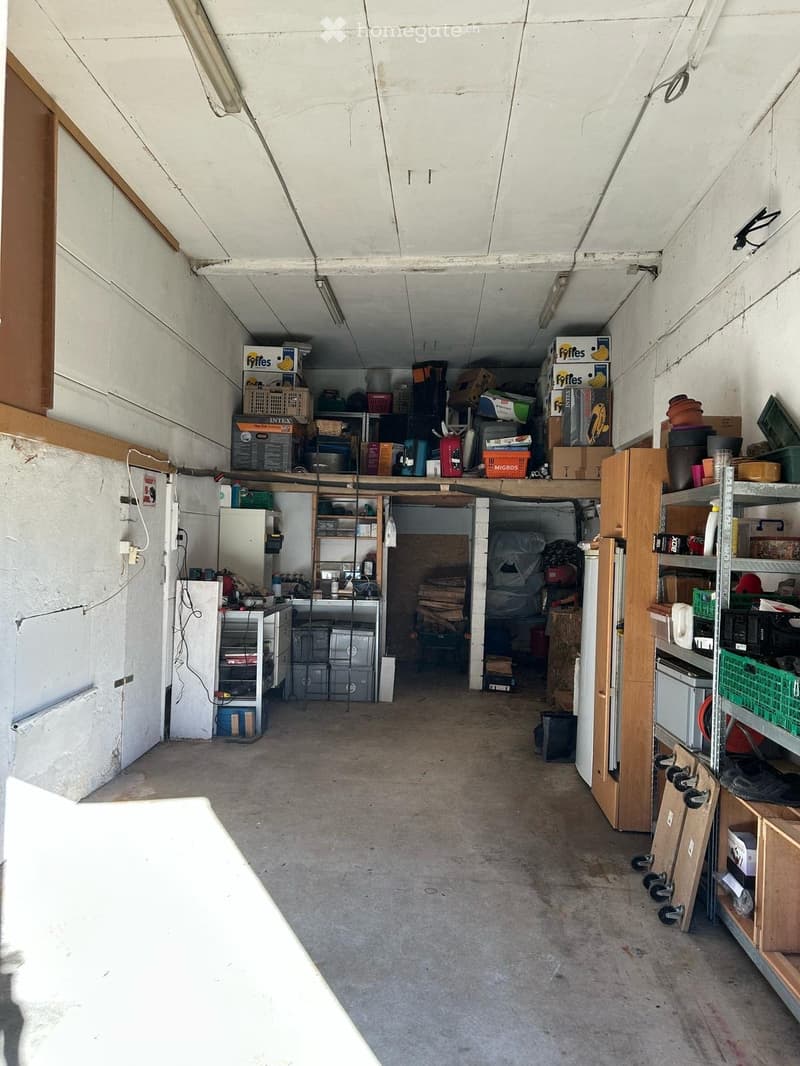 A spacious, unfinished garage with concrete flooring and white walls. Shelves on both sides hold various items including storage bins, tools, and boxes. A refrigerator is positioned against the left wall, and a doorway leads to another storage area in the