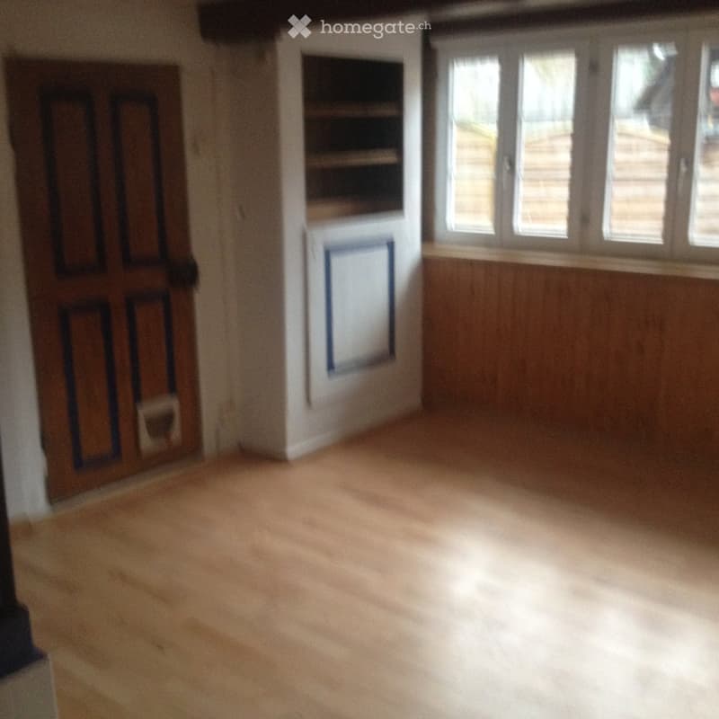 Empty room with wooden flooring, white walls, and large windows allowing natural light. Features a wooden door and a built-in shelf unit.