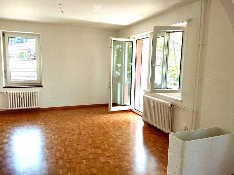 Empty room with wooden flooring, light grey walls, and large windows with open sliding doors leading to a balcony area.