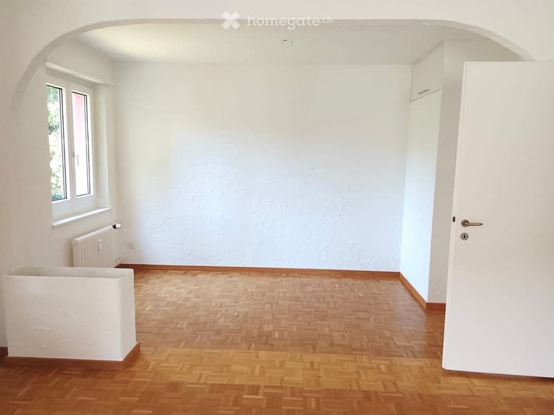 Empty room with wooden herringbone flooring, white walls, a single window with white frame, a radiator, and a white door with a silver handle.