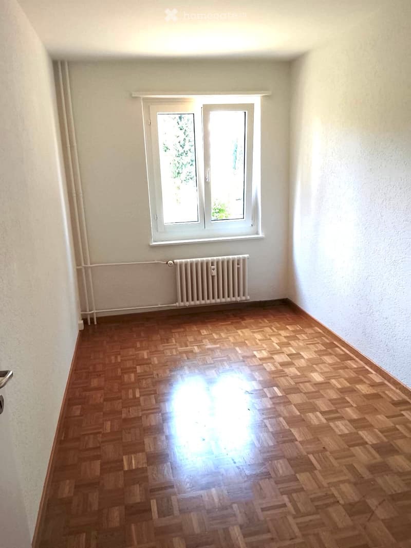 Empty room with wooden parquet flooring, white walls, a window with white frame, and a radiator below the window.