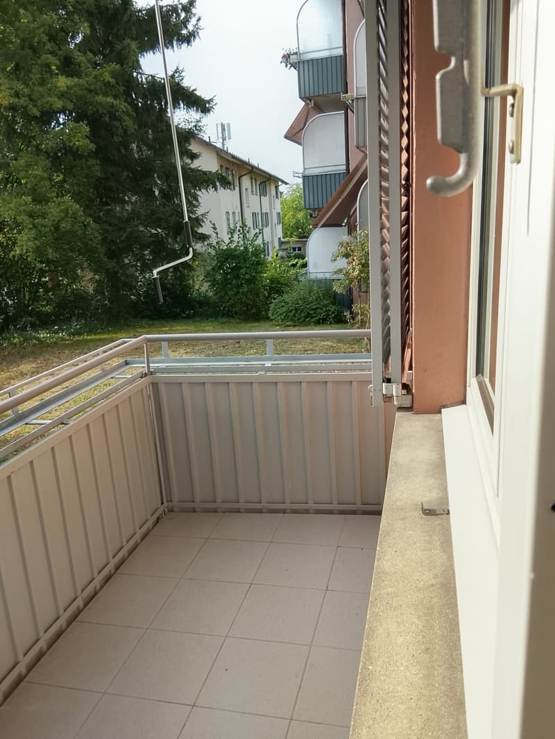 Balcony with tiled floor, metal railing, and view of greenery and neighboring buildings.
