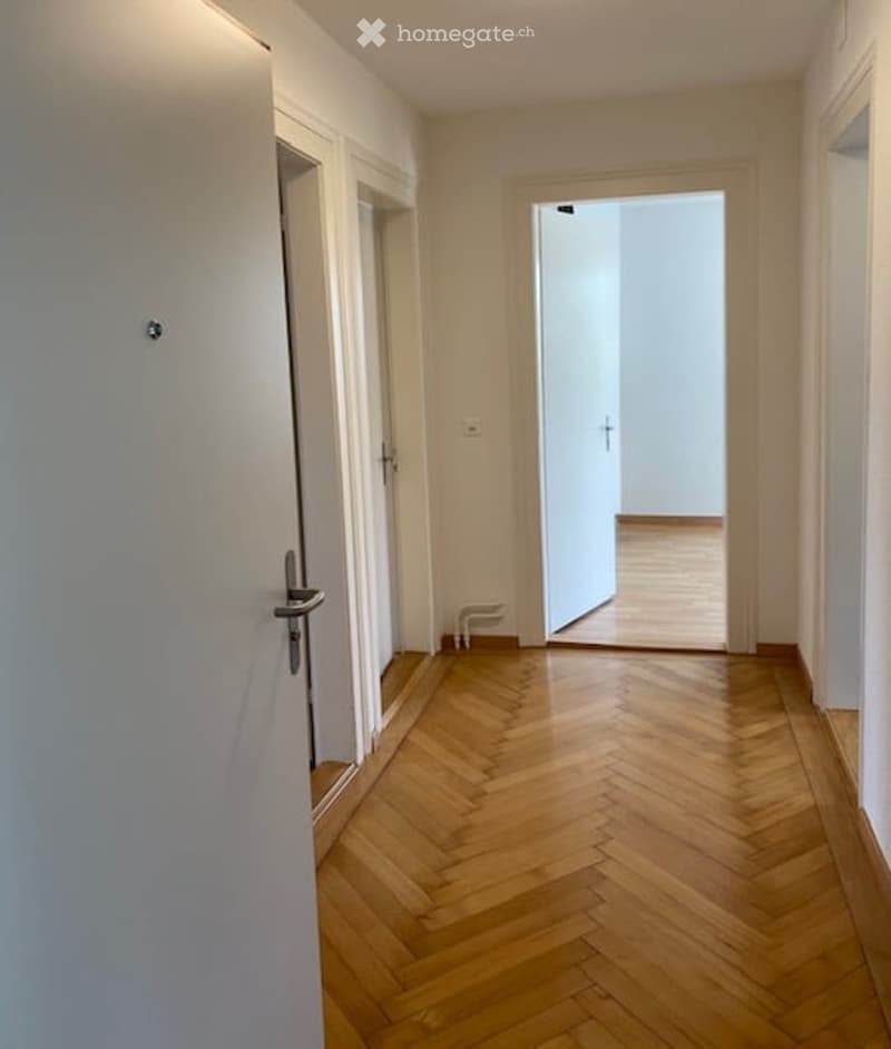 Empty hallway with wooden herringbone flooring, white walls, and open doorway leading to another room.