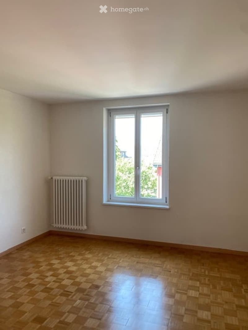 Empty room with wooden flooring, white walls, a single window with white frame, and a radiator on the wall.