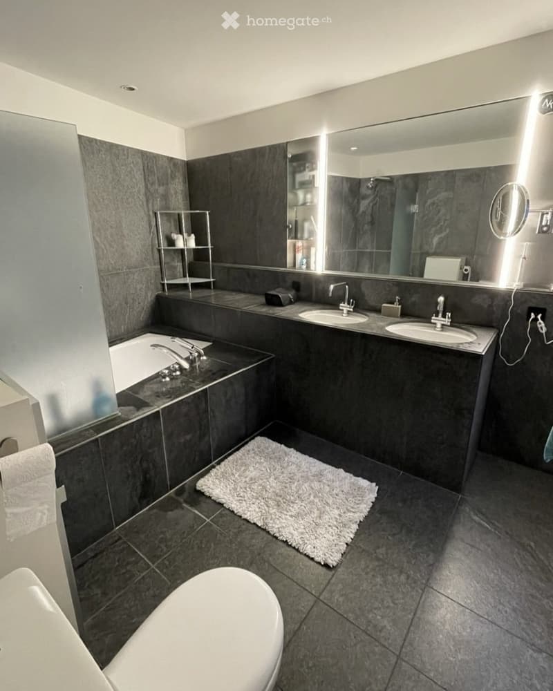 Modern bathroom with dark grey tiles, a bathtub, two sinks, a large mirror, a shelf, and a white rug on the floor.
