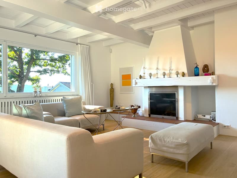 Bright living room with white walls, wooden floor, large window with view, fireplace, sofa, ottoman, coffee table, decorative items on mantel.