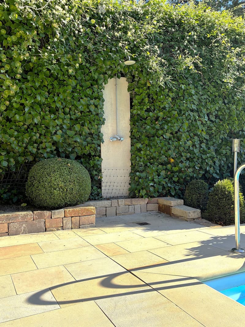 A well-maintained garden area with a swimming pool, surrounded by lush greenery, stone walls, and neatly trimmed shrubs.