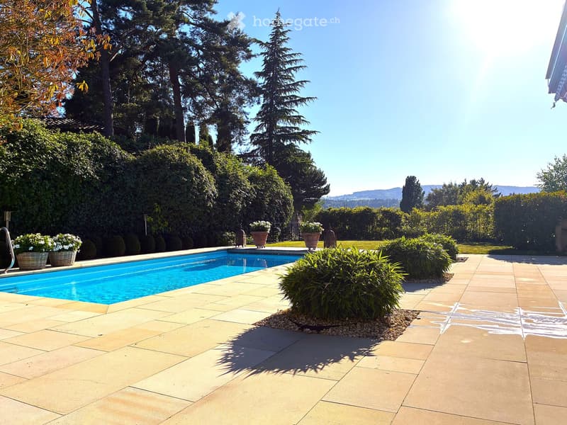 A well-maintained garden area with a swimming pool, surrounded by lush greenery, trees, and neatly arranged potted plants. The pool is surrounded by a tiled patio area, and the background features a scenic view of hills and trees.
