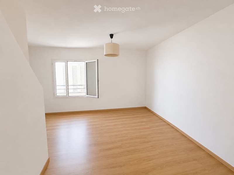 Empty room with wooden flooring, white walls, and a single hanging light fixture. A window with shutters is open, allowing natural light to enter.
