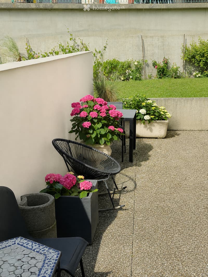 A modern balcony or terrace with a gravel floor, featuring a black wireframe chair, a grey stone pot, and several potted plants with pink and white flowers. The area is enclosed by a concrete wall with a small garden visible beyond.