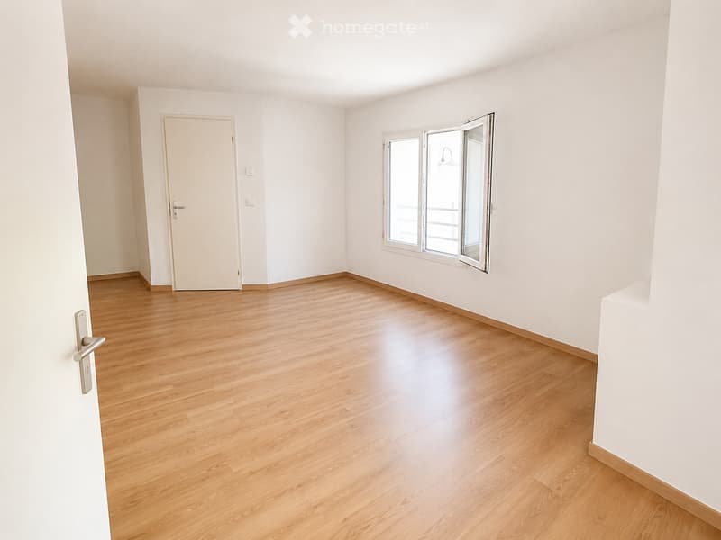 Empty room with light wooden flooring, white walls, a closed door, and a large window with blinds partially open.