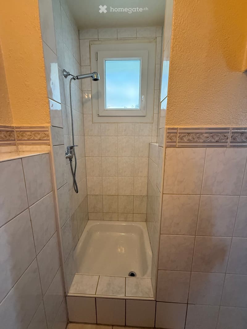 Small bathroom with a bathtub, tiled walls, and a showerhead attached to the wall. A small window is positioned above the bathtub.