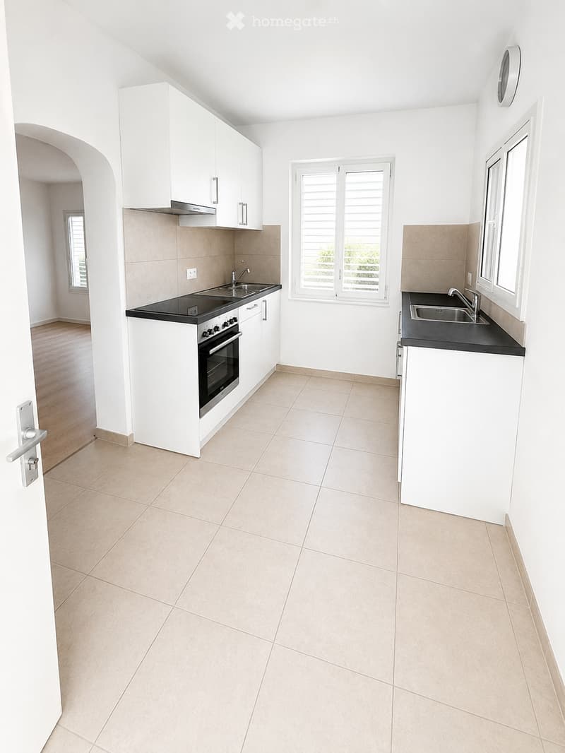Modern kitchen with white cabinets, black countertops, and stainless steel appliances, including an oven and sink. The space features tiled flooring and large windows with blinds.