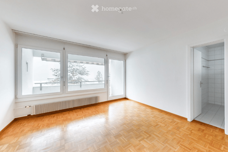 Empty room with wooden flooring, large windows, radiator, and a view of a balcony area.