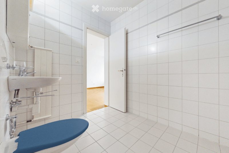 Modern bathroom with white tiled walls and floor, a blue toilet seat, a white sink with chrome faucet, and a door leading to another room.