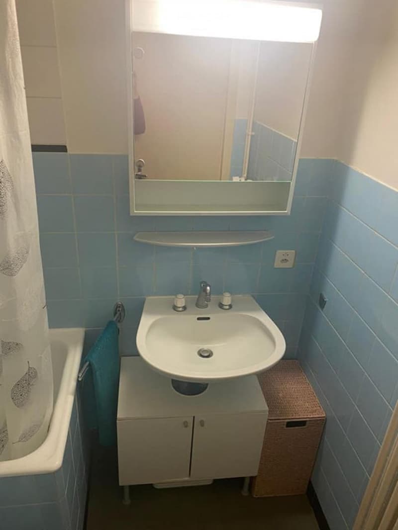 A small bathroom with blue tiled walls, a white sink with a mirror above it, a small cabinet beneath the sink, and a bathtub with a towel hanging on the side.