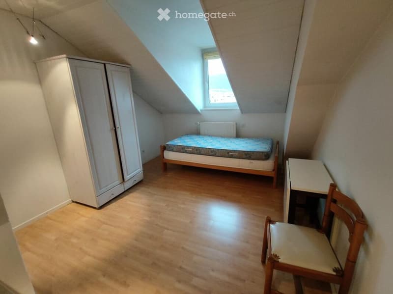 Cozy attic bedroom with wooden flooring, slanted ceilings, and a skylight window. Features a single bed, wardrobe, small table, and chair.