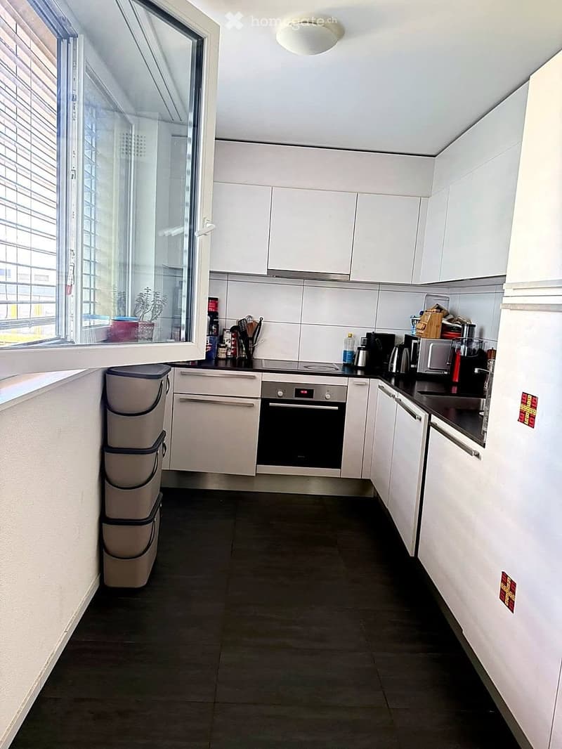 Modern kitchen with white cabinets, black countertops, and stainless steel appliances including an oven, microwave, and refrigerator. Features a large window with blinds and a trash bin on the floor.