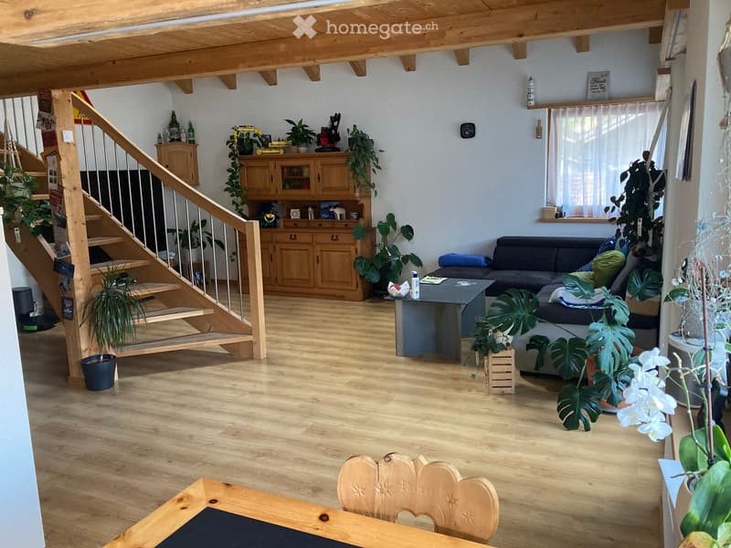 Bright living room with wooden floors, exposed wooden beams, a staircase leading to an upper level, a sofa, coffee table, and a wooden cabinet with glass doors.