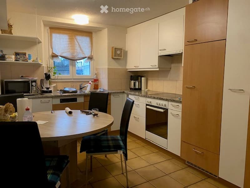 Modern kitchen with white cabinets, tiled floor, and a round dining table with chairs. Appliances include a microwave, coffee maker, and oven. A window with blinds is visible above the sink.
