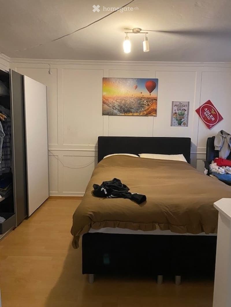 Cozy bedroom with a large bed, wardrobe, and artwork on the wall. The bed is made with a brown duvet and pillows, and there is a pile of clothes on the bed.
