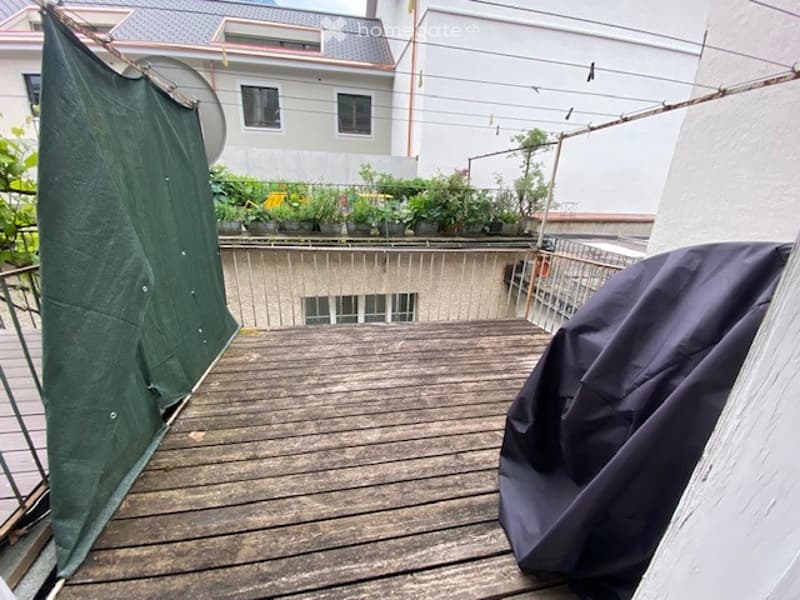 Wooden balcony with metal railing, covered with a green tarp on one side and a dark blue cover on the other, surrounded by potted plants and buildings in the background.