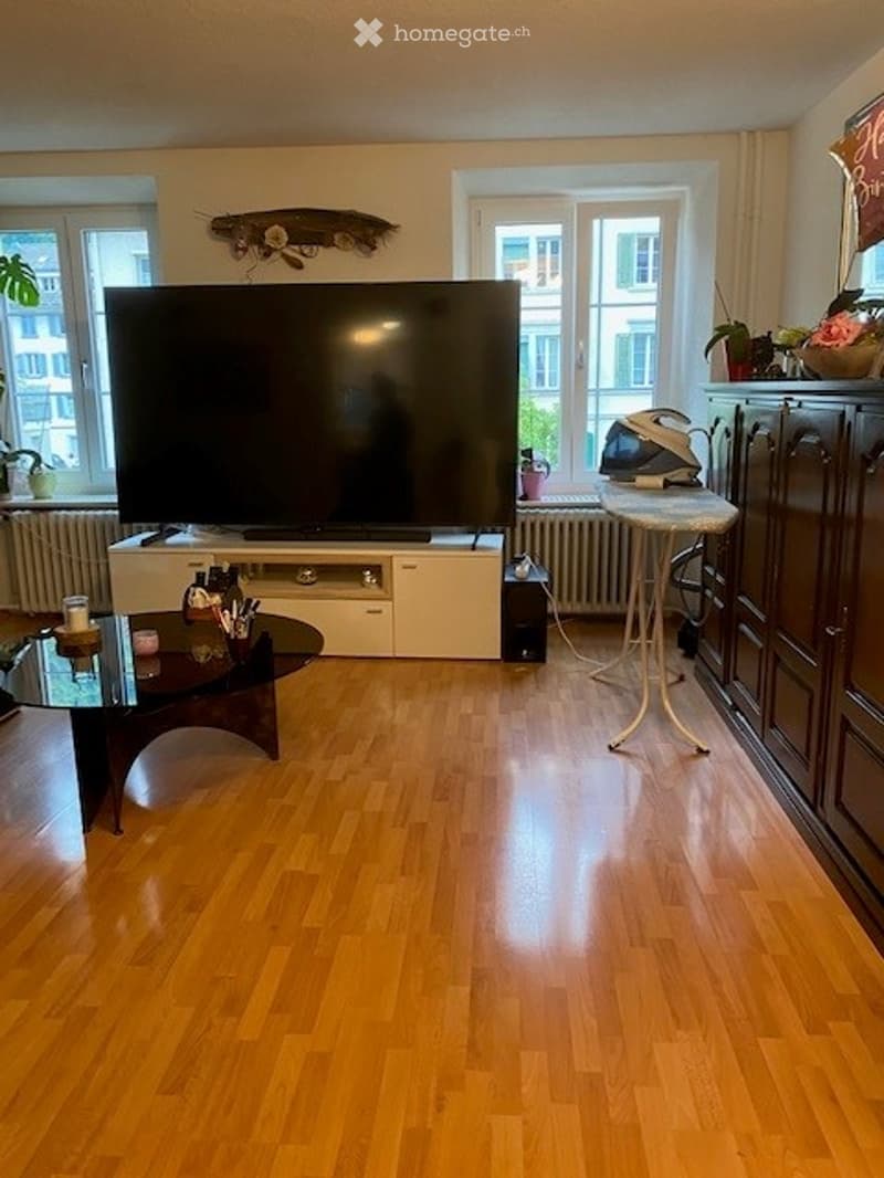 Living room with hardwood flooring, large flat-screen TV on a white stand, glass coffee table, ironing board, and decorative items on a cabinet.