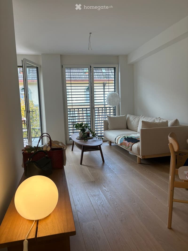 Modern living room with light-colored sofa, coffee table, wooden floor, and large windows with blinds.
