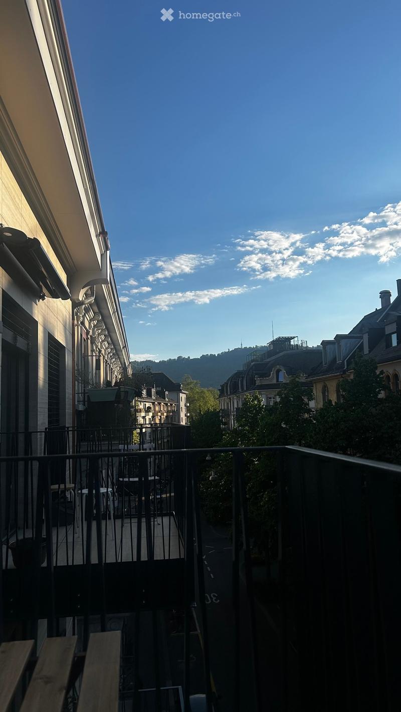 Balcony with metal railing, overlooking a cityscape with buildings, trees, and a distant hill under a clear blue sky.