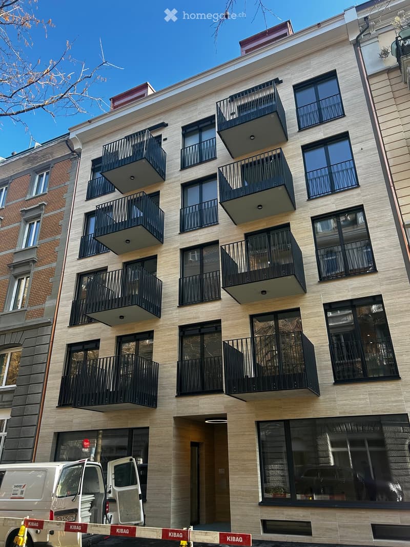 Modern multi-story building with large windows and balconies, featuring a contemporary design with light-colored facade and black railings.