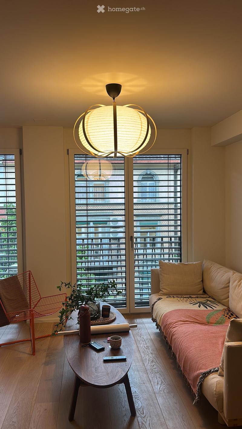 A modern, compact living space with a sofa, coffee table, chair, and a hanging lamp. The room features large windows with a view of a balcony area.